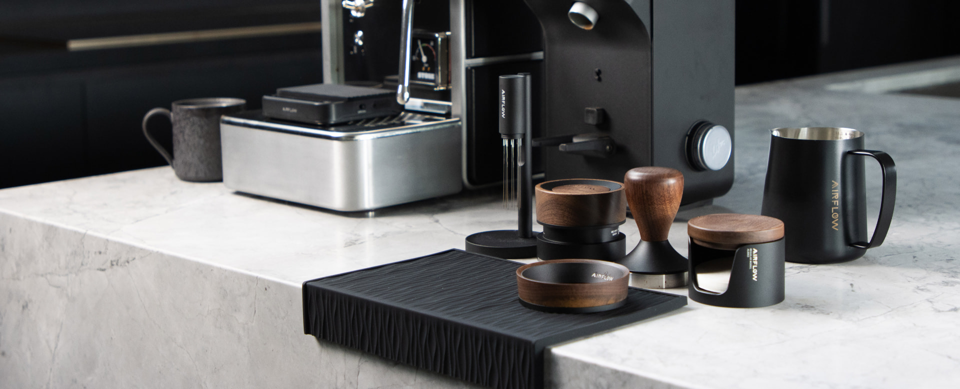 Airflow espresso tools on a marble counter — tampers, dosing tools, jugs and a corner tamping mat in black and walnut.
