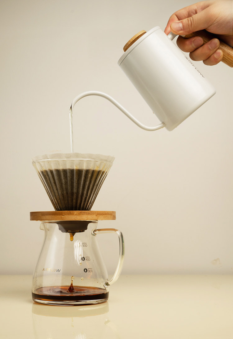 A white Airflow gooseneck kettle pouring water into a glass coffee dripper with a wooden collar.
